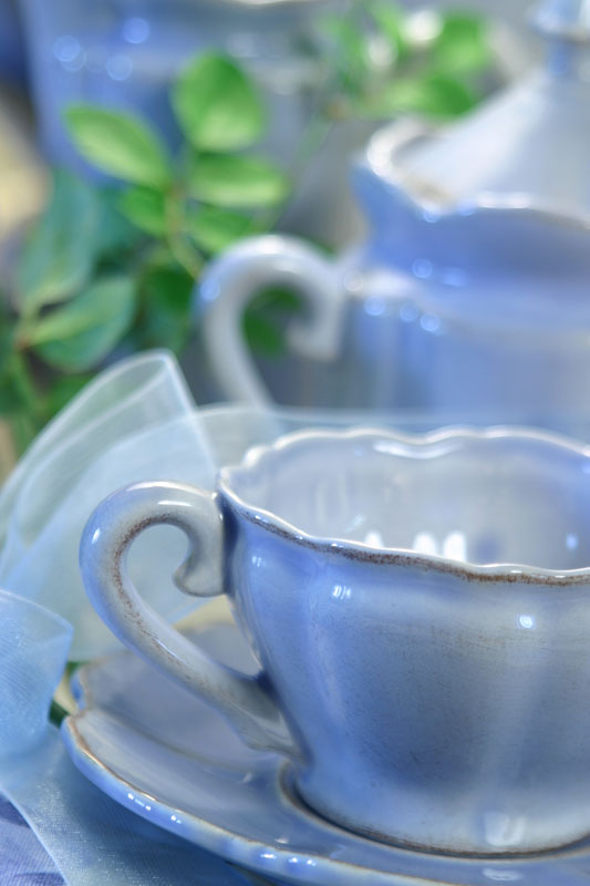 Light blue scalloped teacup and saucer with a matching teapot and green leaves in the background