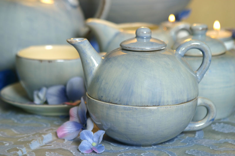 Light blue ceramic tea-for-one set stacked on a table with matching dishes, blue flowers, and lit candles