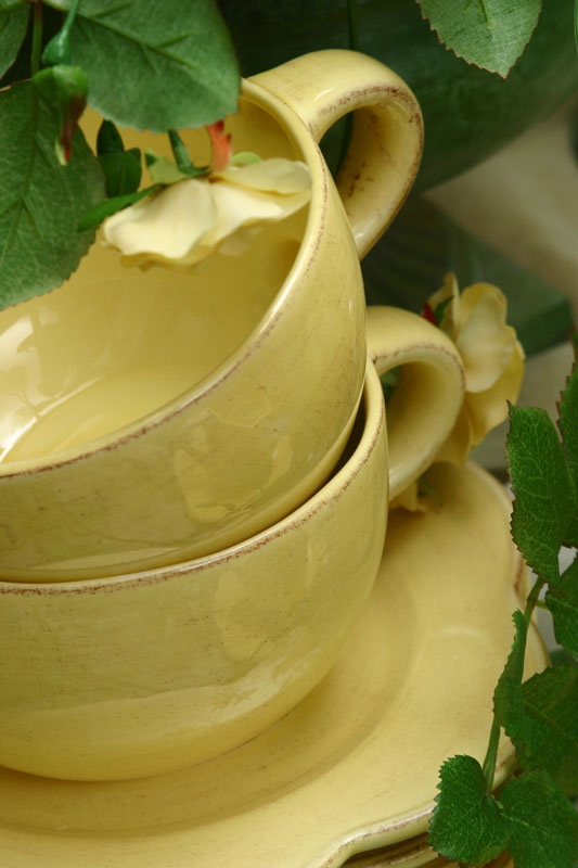 Two yellow ceramic cups stacked on a saucer, surrounded by green leaves and a yellow flower