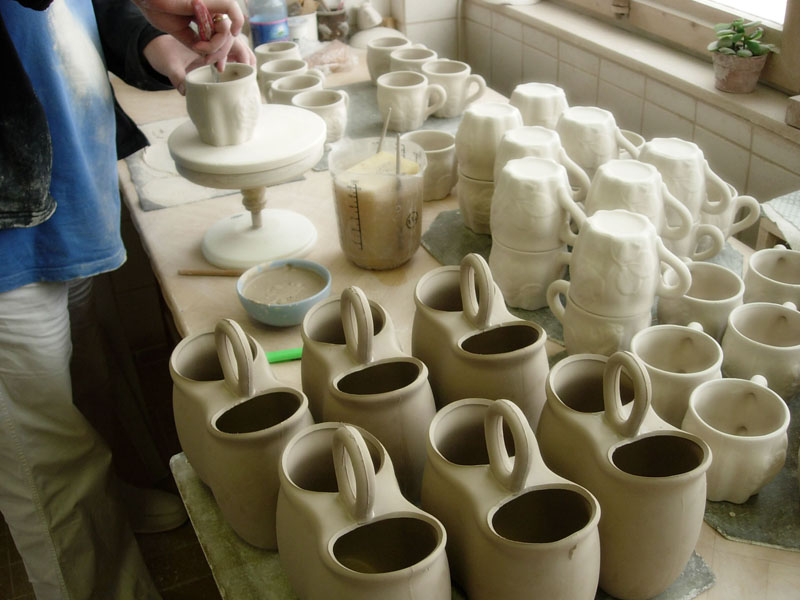 A potter works on a ceramic cup at a workbench filled with many unfinished mugs and double-chambered vessels